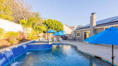 A backyard oasis featuring a pool adorned with blue tiles, complemented by umbrellas and an above-ground spa. Photo