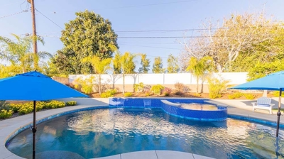A backyard retreat boasting a pool with striking blue tile work, umbrellas for shade, and an above-ground spa. Photo
