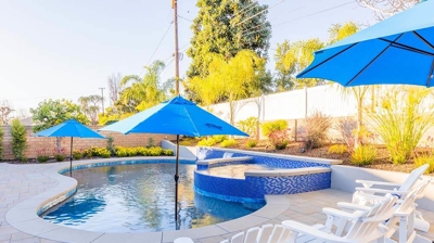 A backyard pool, distinguished by blue tiles, umbrellas, and an above-ground spa. Photo
