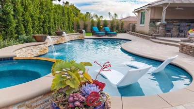 A serene backyard retreat with a pool, lounge chairs, adjacent jacuzzi, and fire pit area enclosed by hedges. Photo