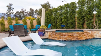 A backyard oasis featuring a swimming pool, white lounge chairs, a jacuzzi, and a fire pit surrounded by hedges. Photo