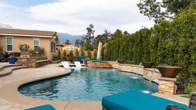 A tranquil backyard sanctuary boasting a pool, white lounge chairs, a jacuzzi, and a fire pit nestled amidst hedges. Photo
