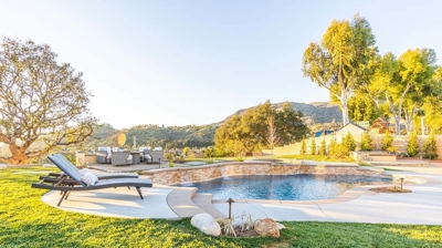 A beautiful backyard oasis with a pool surrounded by grass and greenery next to a fire pit with conversation seating. Photo
