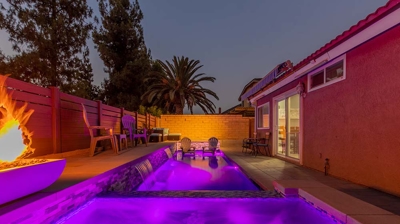 A vast pool with a built-in spa, next to a fire pit and plenty of seating, illuminated by pink lights. Photo