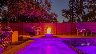 A grand pool with a spa, by a fire pit and ample seating, featuring soft pink pool lights. Photo