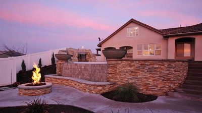 Outdoor fireplace with flame alongside pool and beautiful home. Photo