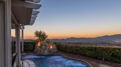 A pool encircled by greenery, adorned by a natural rock waterfall, and illuminated by fire features. Photo