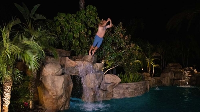 A lit backyard pool surrounded by palm trees and a man jumps off of the top of the waterfall into the pool. Photo