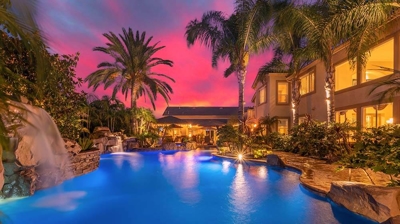 Tranquil oasis surrounded by swaying palms, featuring a pool aglow in blue light against a pink sunset sky. Photo