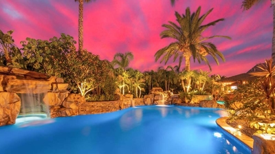 Tropical backyard retreat amidst palm trees, with a shimmering blue pool under a pink sunset. Photo