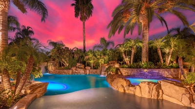 Exotic backyard sanctuary framed by palm trees, offering a tranquil blue pool set against a pink sunset. Photo
