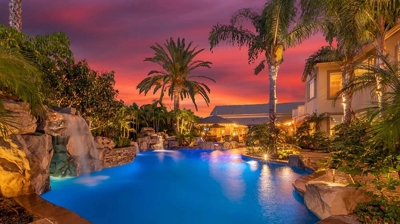 Palm-fringed backyard paradise with a pool illuminated in blue hues, set against a backdrop of a rosy sunset. Photo